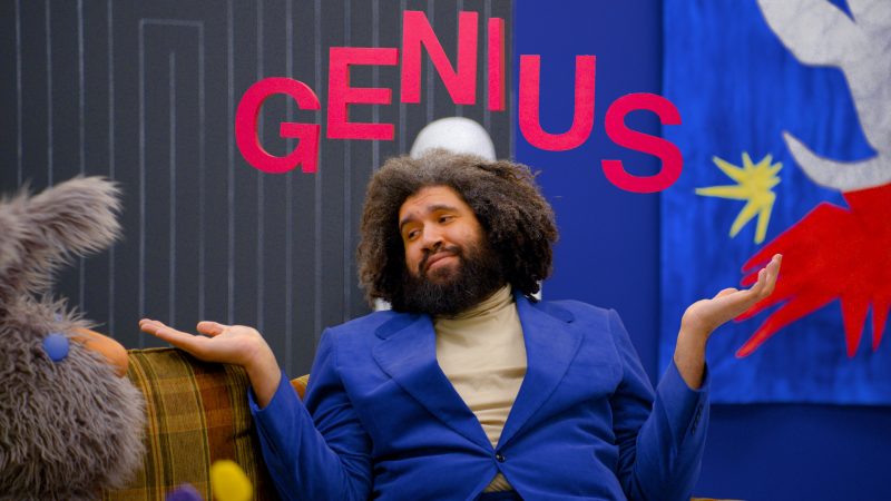 In a still from Jayson Musson's His History of Art, Musson shrugs and looks at his cohost Olli while the word "genius" appears in red letters around his head in a halo.