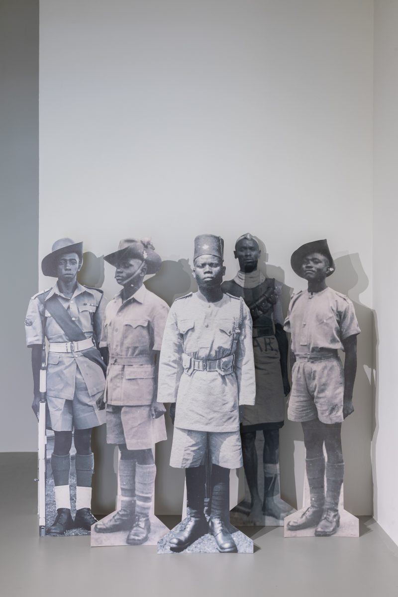 A photo shows a group of five African male cut out figures standing together in a corner of a room, all are wearing military uniforms and at attention.