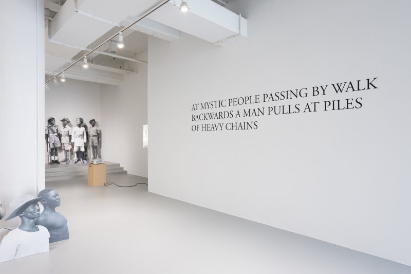 A photo shows a gallery installed with several black and white standing cutout figures, some in background up a set of stairs and some in foreground appearing to look at a wall on which there is writing.
