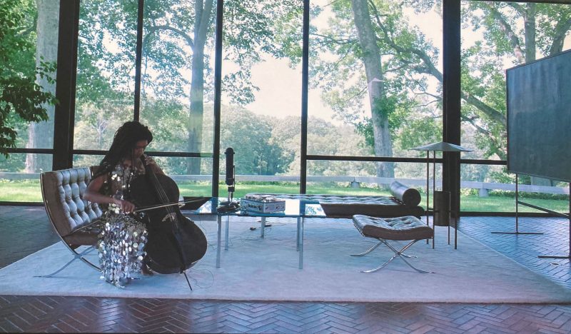 A color photo shows a woman dressed in a silver-sparkly dress sitting alone on a couch in a “glass house” with beautiful trees outside the windows and playing a cello.