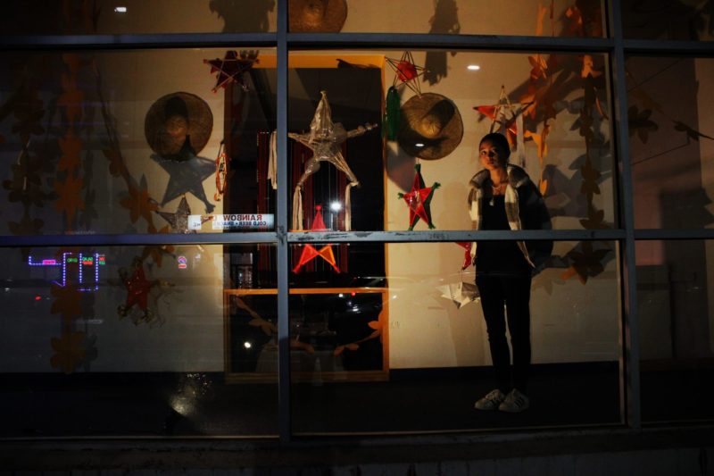 A large shop window seen at night lit up with warm yellow lights shows an art installation of many hand-made perhaps paper or fabric stars in many colors and sizes suspended in the space and several woven straw hats on the back wall around a dark doorway. A young woman stands amidst the stars looking slightly wary at something outside the window.