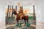 Life-sized sculpture depicting an evolved form of future humans wearing intricately patterned orange and brown clothing, riding a horse in front of a hand-painted forest environment with sparse trees and an orange sky.