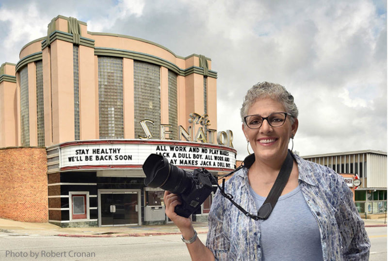 Portrait of Baltimore photographer Amy Davis smiling with a camera in front of one of their subjects, a closed movie theater called the Senator.