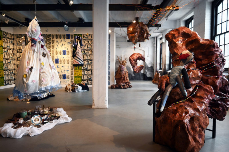 A colorful photo shows an installation of art by two artists, on the left, a dress, wallpaper and an historic artifacts on cloth on the floor and on the right a number of large, shiny brown sculptural objects that suggest the abstracted human body in various poses suggesting discomfort or even torture.
