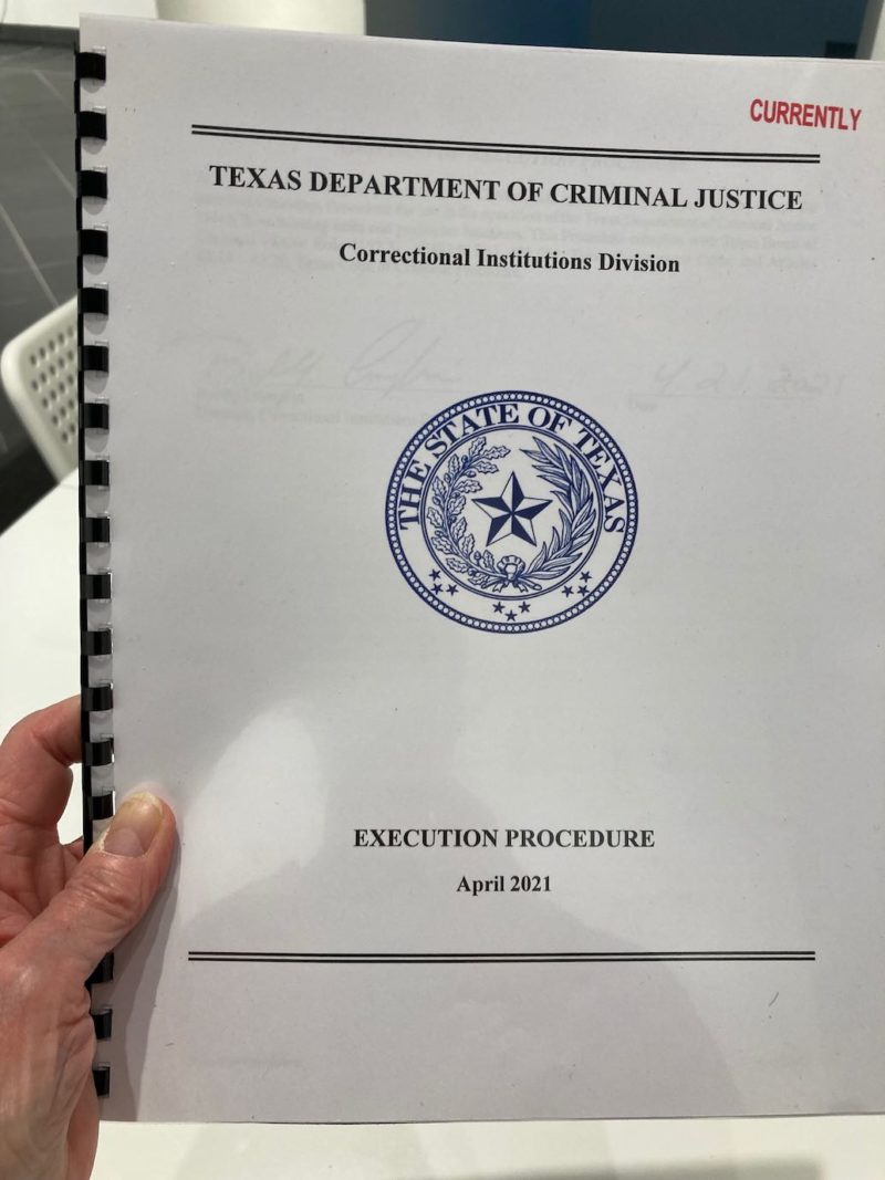 A color photo shows a hand holding a copy of a training manual that is spiral bound at the left and whose cover indicates it is the Texas Department of Criminal Justice Correctional Institutions Divisions Execution Procedure manual from April, 2021. Red letters at the top mark it “CURRENTLY” meaning currently in use.