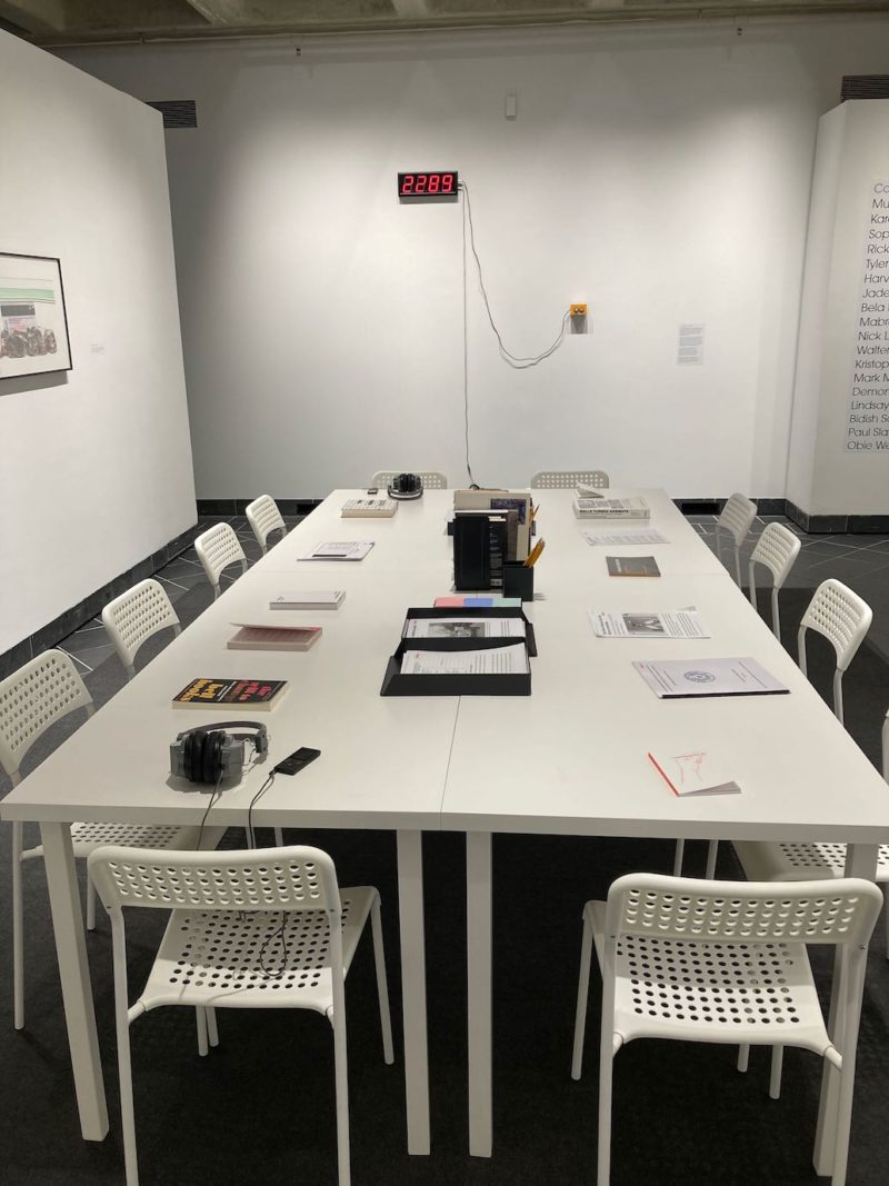 A color photo shows a room with no windows and a long white table in the middle with many white chairs around it, with books and papers on the table and on the far wall is a digital display reading “2289” in red letters, referring to the number of prisoners currently on death row in the US.