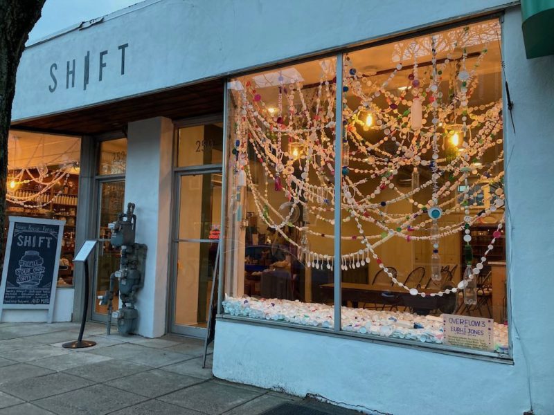 A color photo shows the exterior of a store in Narberth PA in a blue-tinted building with the store’s name “Shift” appearing above the main entrance. In the window on the right is an art installation, made of colorful ribbon-like strands of plastic spoons, beads and other small plastic wares. In the window shelf are hundreds of white and colored plastic bottle caps. The label in the window says the work is “Overflow3” by Eurhi Jones.