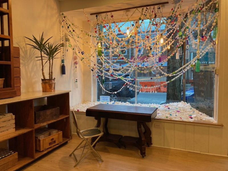 A color photo shows the interior of a store, Shift” located in Narberth, PA. In the window is an art installation, made of colorful ribbon-like strands of plastic spoons, beads and other small plastic wares. In the window shelf are hundreds of white and colored plastic bottle caps.