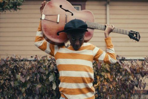 A black man in an orange and white shirt and black hat holds a dark wooded guitar outside looking up at the camera.
