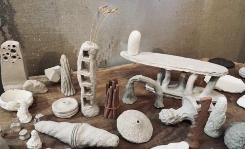 white clay objects of varying shapes and forms sit atop a greyish piece of wood. The objects take on obelisk forms with holes or squares cut through, disks, bowls, stone like shapes. All cluttering next to one another.