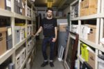 Portrait of artist Rami George in the John J. Wilcox Jr. Archives at the William Way LGBT Community Center, wearing cuffed jeans and a black t shirt and leaning a hand on the archive shelf.