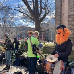A photo shows a beautiful, sunny day on Temple University’s campus, with wispy clouds in the sky, and in the foreground, a band, including guitar, saxophone and trumpet, and featuring a drummer who is wearing a Gritty mask on his head. They are playing for the graduate student union strike protest on campus.