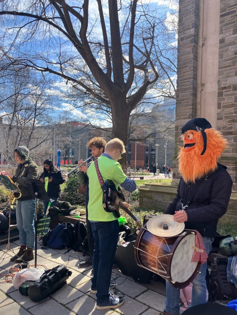 A photo shows a beautiful, sunny day on Temple University’s campus, with wispy clouds in the sky, and in the foreground, a band, including guitar, saxophone and trumpet, and featuring a drummer who is wearing a Gritty mask on his head. They are playing for the graduate student union strike protest on campus.
