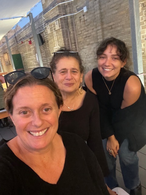 A color photo shows a three women, smiling, all wearing black, taking a selfie in a hallway that is brightly lit and welcoming. 