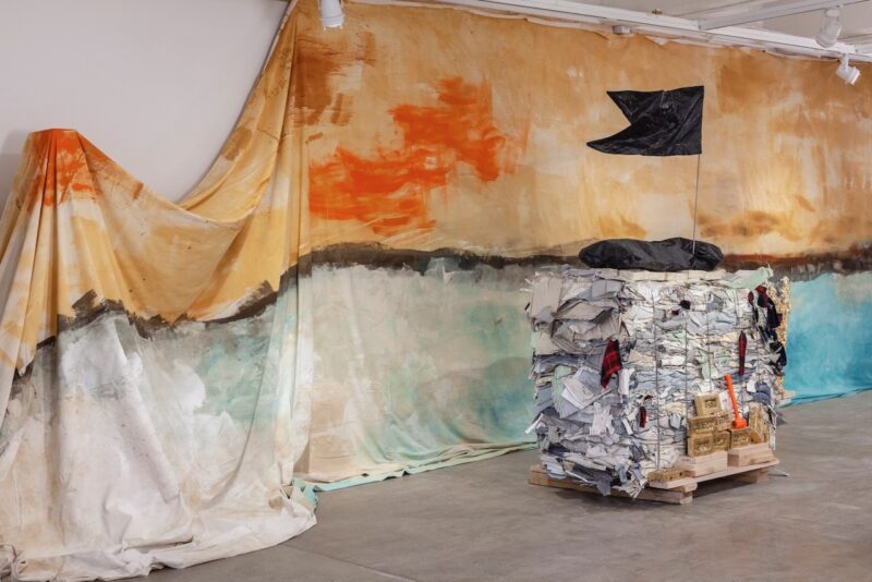 A color photo shows an art installation by Henry Taylor in a high-ceilinged gallery, where, on the floor is a wood pallet with a large bundle of grey and white flattened discarded objects held down by metal ropes, atop of which is a black plastic bag, perhaps a body bag, which is holding up a stark black flag on a pole, and in the background, a brushy painted theatrical sea scape on a large sheet of fabric, with the sky a roistering ochre color with red blotches suggesting clouds touched by sunset and the bottom of the cloth painted aqua suggesting the sea with waves pounding a retaining wall, and thus, the bundle might be like a ship on the water, its black flag signaling piracy.