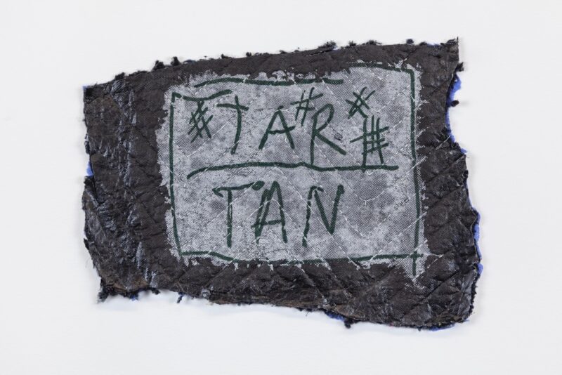 A photo of a black quilted moving blanket with its edges tattered and its shape asymmetrical has been drawn on, with a think coat of white paint in a square in the middle with the words “TAR” and ‘TAN” and some # signs drawn on top in black marker, with black lines encircling the drawn square and a black line separating ‘TAR” on the top from ‘TAN” on the bottom.