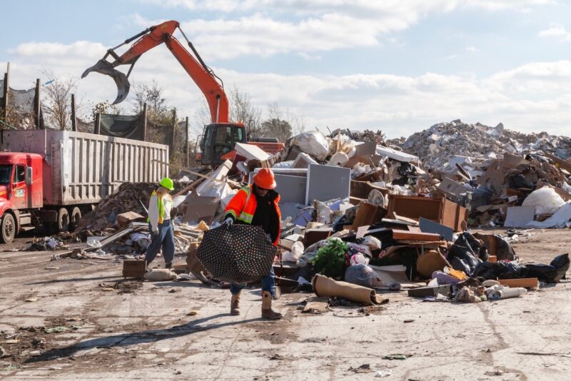 Two people scavenge materials from a recycling center, one in the red hard hat and jacket is artist Henry Taylor, and the other in the green hardhat and green vest is Abby Lutz of the Fabric Workshop and Museum. They are gathering materials for Taylor’s exhibit at the FWM. In the background is a dump truck on the left and an orange backhoe, and there is a big pile of debris on the concrete surface.