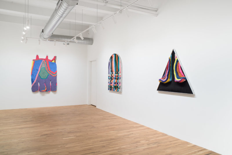 A color photo shows three large soft-sculptural objects on the walls of a high-ceilinged gallery with a light wood floor. The objects are shaped (l-r) like a limp hand hanging, a rounded-top window and a triangle and they all have bright colors and suggest abstracted landscapes interpreted through a domestic lens.