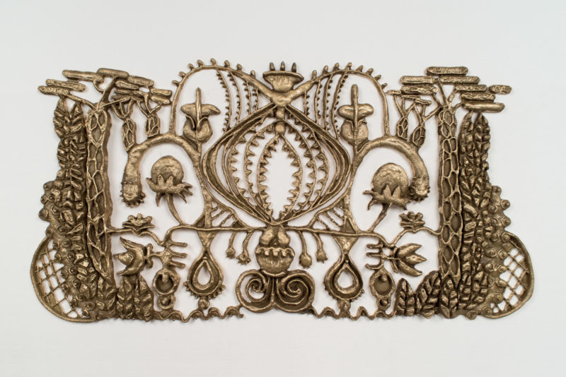 A color photo of a large brass wall adornment featuring curlicues, seed pods, seeds, nets, and a central image suggesting an onion, all of the elements touching and connected as if from a kind of baroque, branching vegetable tree of life.