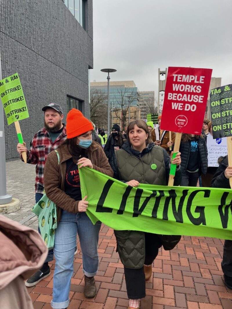 A photo shows a gray, cloudy day on Temple University’s campus, and in the foreground, Temple graduate students and supporters are marching, carrying bright green and red picket signs and a green cloth banner with messages in support of the striking graduate students.