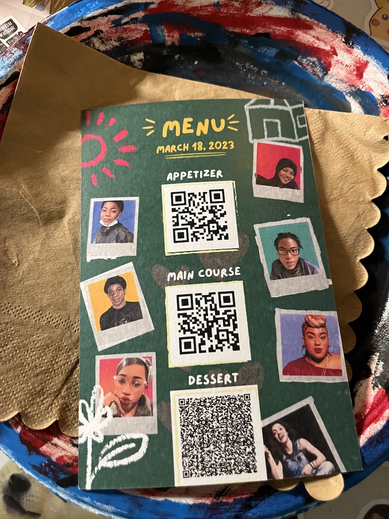 A colorful photo shows a printed program card with QR codes in a column in the middle and seven portrait faces in two columns flanking the QR codes. The program sits on top of a beige paper napkin which is on top of a red-white-blue painted plate.