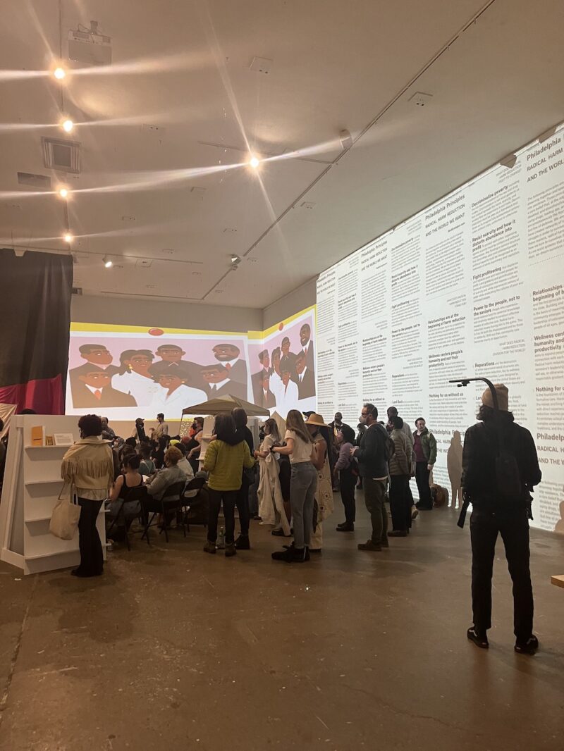 A large crowd is shown gathering for a workshop Rosine 2.0, in a high-ceilinged art space in Philadelphia, with all the people looking to a far wall, perhaps at the projected videos in the back walls or perhaps listening to a speaker.