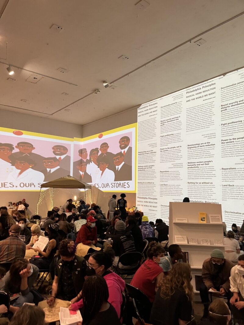 A photo shows a crowd sitting at tables in a workshop at Rosine 2.0 at the Icebox Project Space, a large, high-ceiling arts venue in Philadelphia. The background shows the people are sitting in a corner of the space, with video projections spanning the corner onto the two back walls, showing a painting of black and brown people together framed in a yellow frame and to the right words about harm reduction.