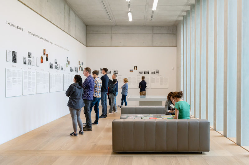 A color photograph shows a number of people studying somedense written and photographic material on two walls in a bright-lit room in a Swiss museum in Zurich. 