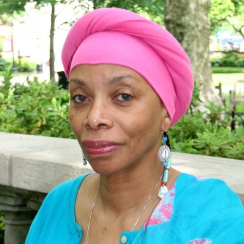 A light skinned black woman in a pink headwrap and blue shirt is pictured from the shoulders up ina nature setting with stone barrier.