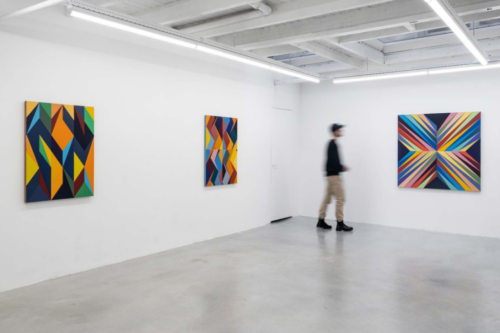 In an white gallery with a concrete floor a blurred man inspects the multicolored angular compositions of Odili Donald Odita.