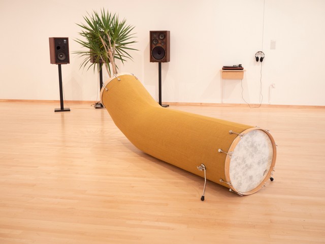 A gallery installation shows an altered drum on the floor in the foreground with a Yucca plant on a stand surrounded by two standing speakers in the background