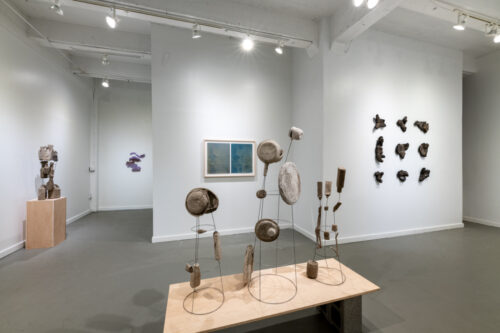 The white gallery space of Tiger Strikes Asteroid a series of three garden wire strructures hold plate like shapes and bottles, another sculpture of a similar beige color pallet is on a pedestal on the left. A purple wall hanging is in the far background, a blue diptych sits behind the closest sculpture. A wall of masks continues on through the right side.