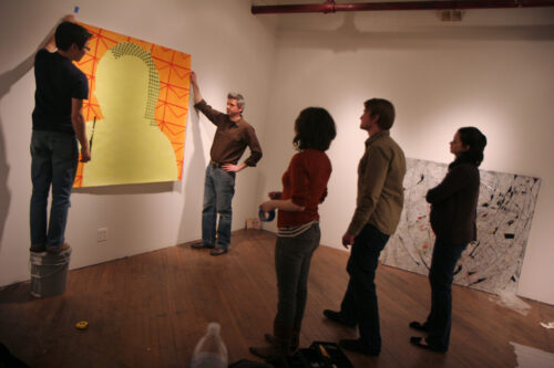 A group of two women and a man stand back and look as two men hang an orange and yellow portrait on a white gallery wall.