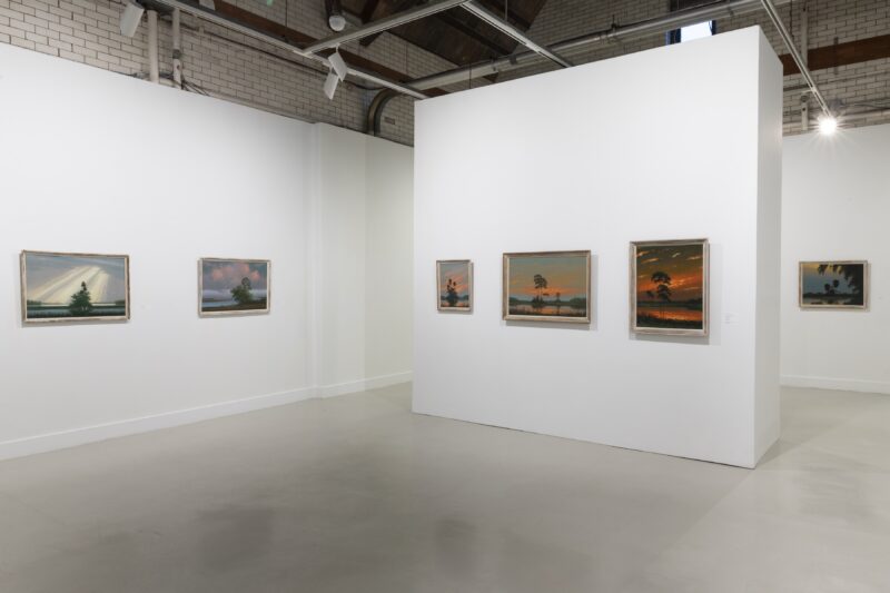 An art gallery with a high-ceiling and polished concrete floor shows three walls on which six landscape paintings sit. There are more paintings in the show but they are not in view in this cropped detail of the gallery.