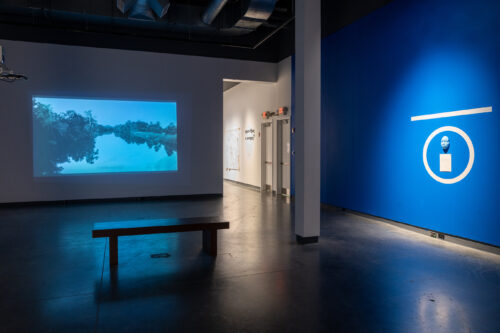 A gallery space with a wood bench facing towards a screen with a river scene. To the right is a blue wall with a white o and flat horizontal line above, in the circle is a blue face.