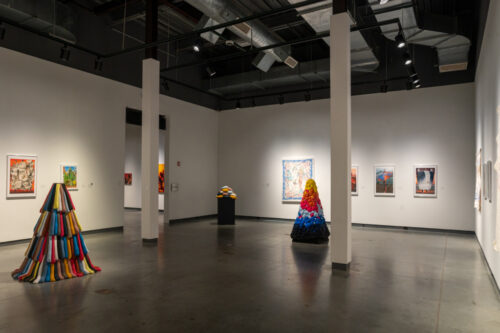 Gallery space with multicolored pyramids of cloth in cylinders, spheres, and rounded pancakes. The walls feature canvases.