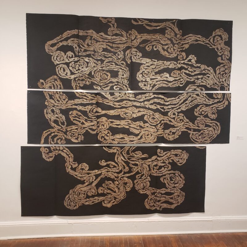 A three-panel triptych shows a meandering, intestine-like or map-like squiggle, colored beige, and sitting on top of a black paper background.