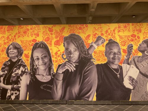 A photo shows a series of wall sized photographic images of five black women over a floral wallpaper in high contrast yellow and orange. The women left to right include a woman dressed in a camouflage jacket looking up, a woman with braids looking straight out and smiling deeply; a woman with twists in her hair holding a pencil in her hand which rests under her chin, she also looks straight out but is straight faced, the next woman holds her fist in the air with a look of joyful resistance on her face; the last woman faces to the left and looks up as she speaks into a microphone.