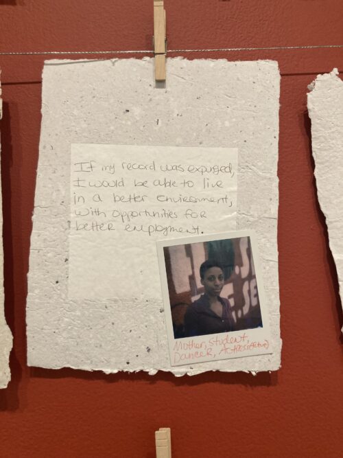 A photo shows a piece of handmade paper clothes pinned to a wire on a red-brown wall, the paper has a note collaged onto it that reads: If my record was expunged, I would be able. To live in a better environment, with opportunities for better employment. On top of this note is a Polaroid photograph of a black woman with writing at the bottom that reads Mother, Student, Dancer, Actress (future). The woman stares out at us with an ambiguous smile.