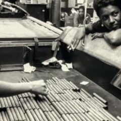 A black woman with short hair with a cigarette loosely hanging from her hand stands in the right middle plane of the picture watches as a boy and man with only their bodies in the shot stock a crate with what appear to be cigars.