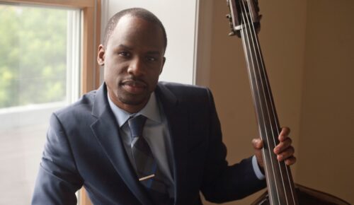 A sharply dressed Black man holds a double bass in his right hand near a window.