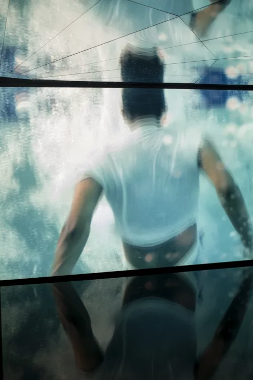 A photo shows a still image of a video on two wall size screens and reflected in a series of mirrors on the ceiling, a portion of a the video is split in two and a shiny black floor resting under it, the screen has an image of a person shown from the back who looks to be jumping into highly reflected water, this image is mirrored above in a fractured screen.