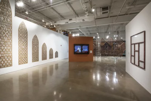 A photo shows a gallery room with artworks hung on opposite walls and one in the center which stands in front of another further back, each object is made of wood. On the right is a window-like structure with panes of glass reflecting the gallery lights, across on the left is a series of six of mashrabiya structures that are square at the bottom and pointed at the top, these range in size from biggest in the front to smallest in the rear and each has a similar radiating pattern within its frame; the objects in the center of the room appears to be a window with a figure sitting within it, the structure behind this object is a series of lattice work squares arrange in a patter creating a wall like structure.