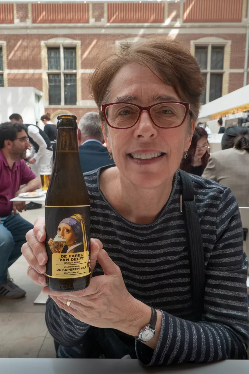 Image of a woman with glasses and a striped shirt holding a beer in a corwded area. The beer features Vermeer's Girl with a Pearl earring drinking out of a stein.