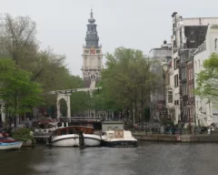 A photo of a river in Amsterdam alongside rows of houses, trees, and a tower daunting in the background.