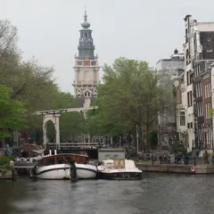 A photo of a river in Amsterdam alongside rows of houses, trees, and a tower daunting in the background.