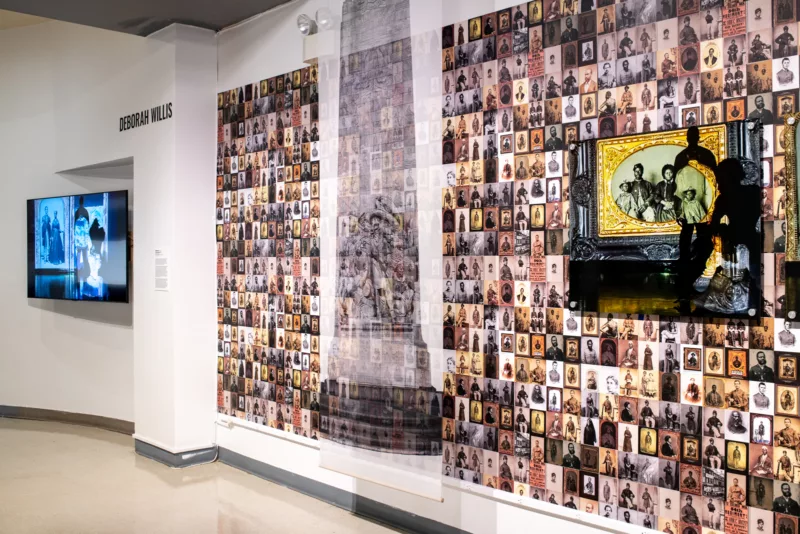 A photo shows a wall covered with photographs that look like they are all from the 19th century, all sepia tones and black and white with oval or ornamental frames around each. Over this wall are two objects, one is an enlarged image of a daguerreotype of a black family and a smaller image of a blurry woman with silhouettes of black figures over and next to the daguerreotype. next to this in the center of the wall is a translucent scrim with a decorative column printed on it. Next to the wall of photographs there is a flat screen with a cyanotype blue images of black figures on the screen.