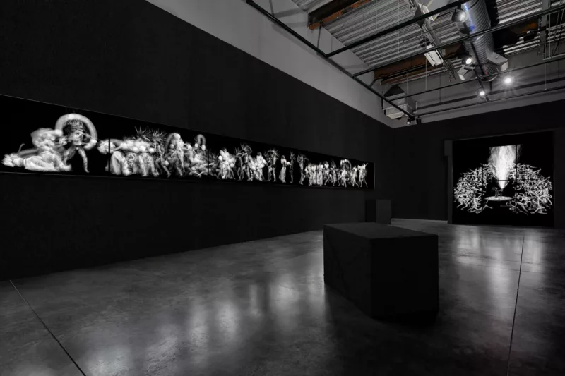 A concrete floored, black walled gallery displaying the OLED electronic canvases of photography John Singletary. The view shows the work Anahata, a horizontal stretch of OLED canvases stretches showing a fantastical black and white image of human like creatures. On the right is another group of canvas in a large square showing humans piling atop each other in space around a central flame creating a ring.