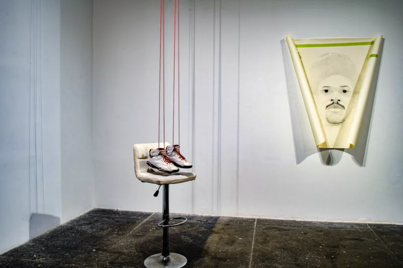 A corner of a gallery with a dark concrete floor shows two pieces of art, one a sculpture with a bar stool, a pair of white leather sneakers and the other, a portrait in black pencil on white canvas, of a Black man with a mustache looking out at you from a V-shaped, curled canvas or paper. 