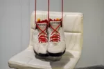 A photo shows a closeup of white Air Jordan’s with red shoelaces, that hover above a white leather bar stool. The red shoelaces disappear upwards giving them the appearance of levitating through magic.